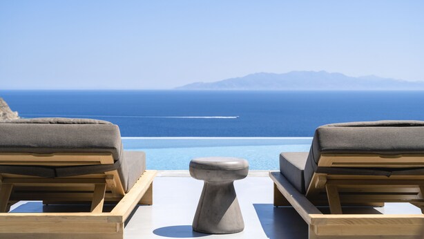 Une vue depuis un balcon, avec deux fauteuils de jardin en bois et une table en pierre, donnant sur une mer bleue et limpide et sur des montagnes lointaines.