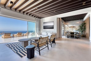 Een ruime woonkamer met een groot raam met uitzicht op de oceaan, een eethoek met een tafel en stoelen, en een gezellige zithoek met kussens en een bijzettafeltje.