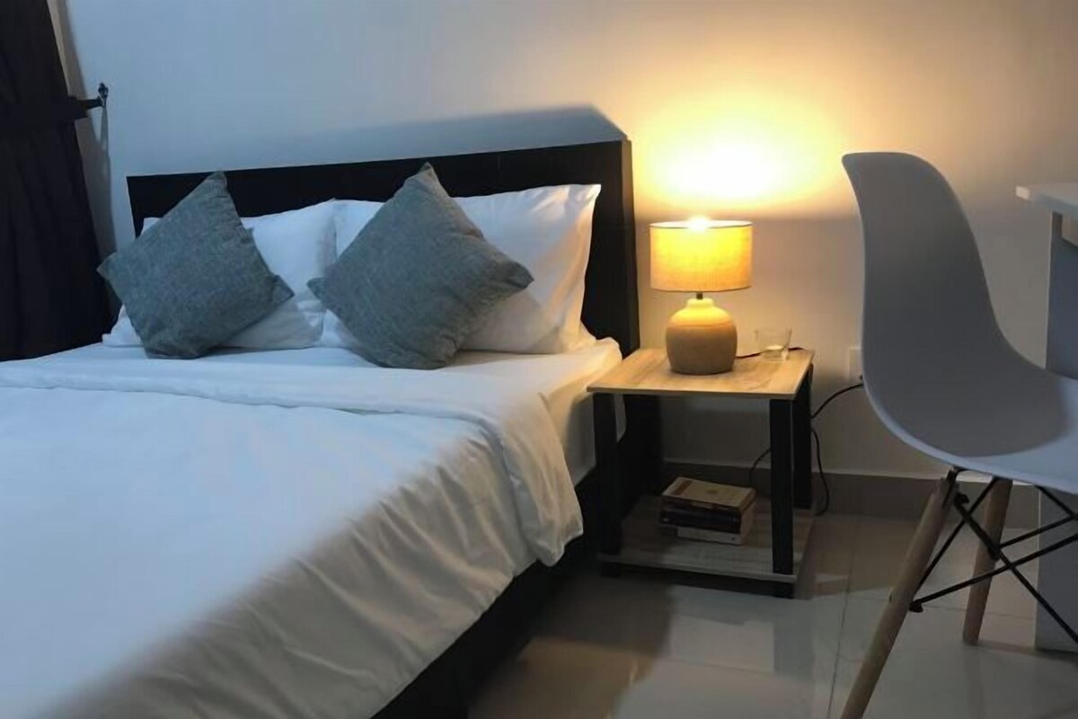 Un lit bien fait, avec du linge de lit blanc et deux oreillers gris, une table de chevet avec une lampe et une chaise moderne.