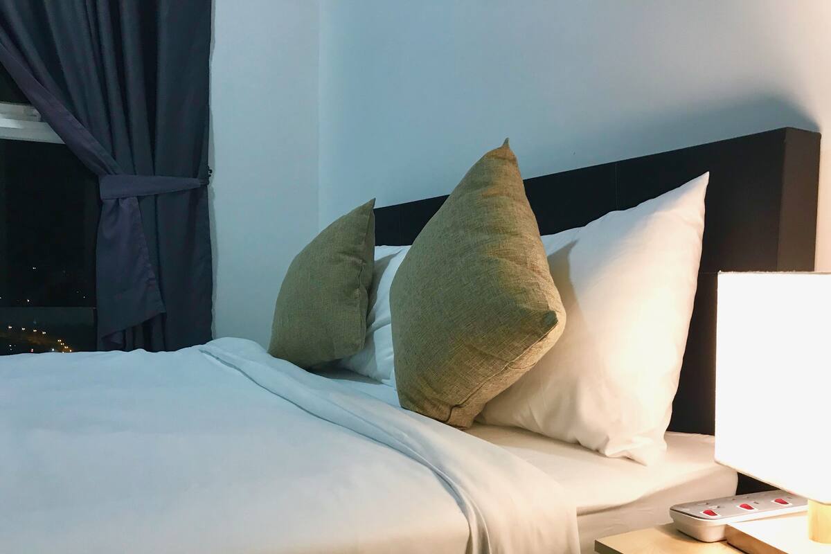 Una cama bien tendida con sábanas blancas y dos almohadas grandes color beige, una mesita de noche con una lámpara, y una ventana con cortinas.