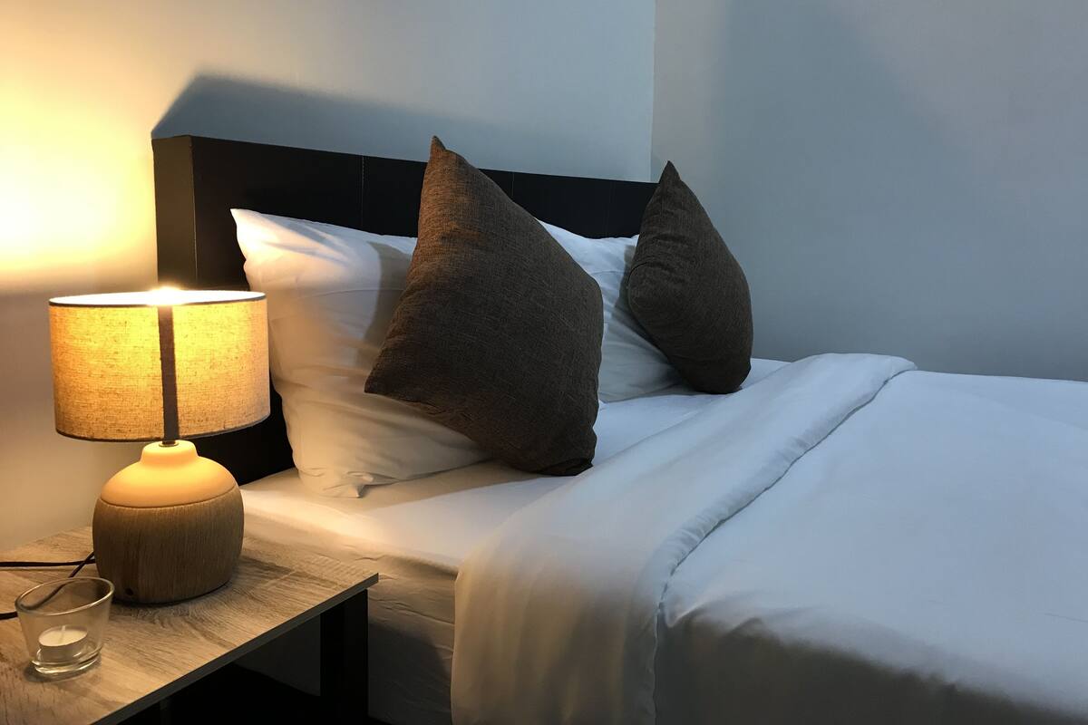 Una cama bien tendida con sábanas blancas, dos almohadas grandes, una mesita de noche con una lámpara y un vaso de agua.