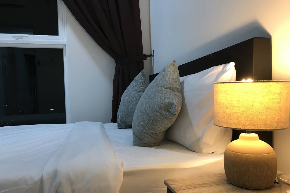Una cama bien tendida con sábanas blancas, dos almohadas grises y una mesita de noche con una lámpara.
