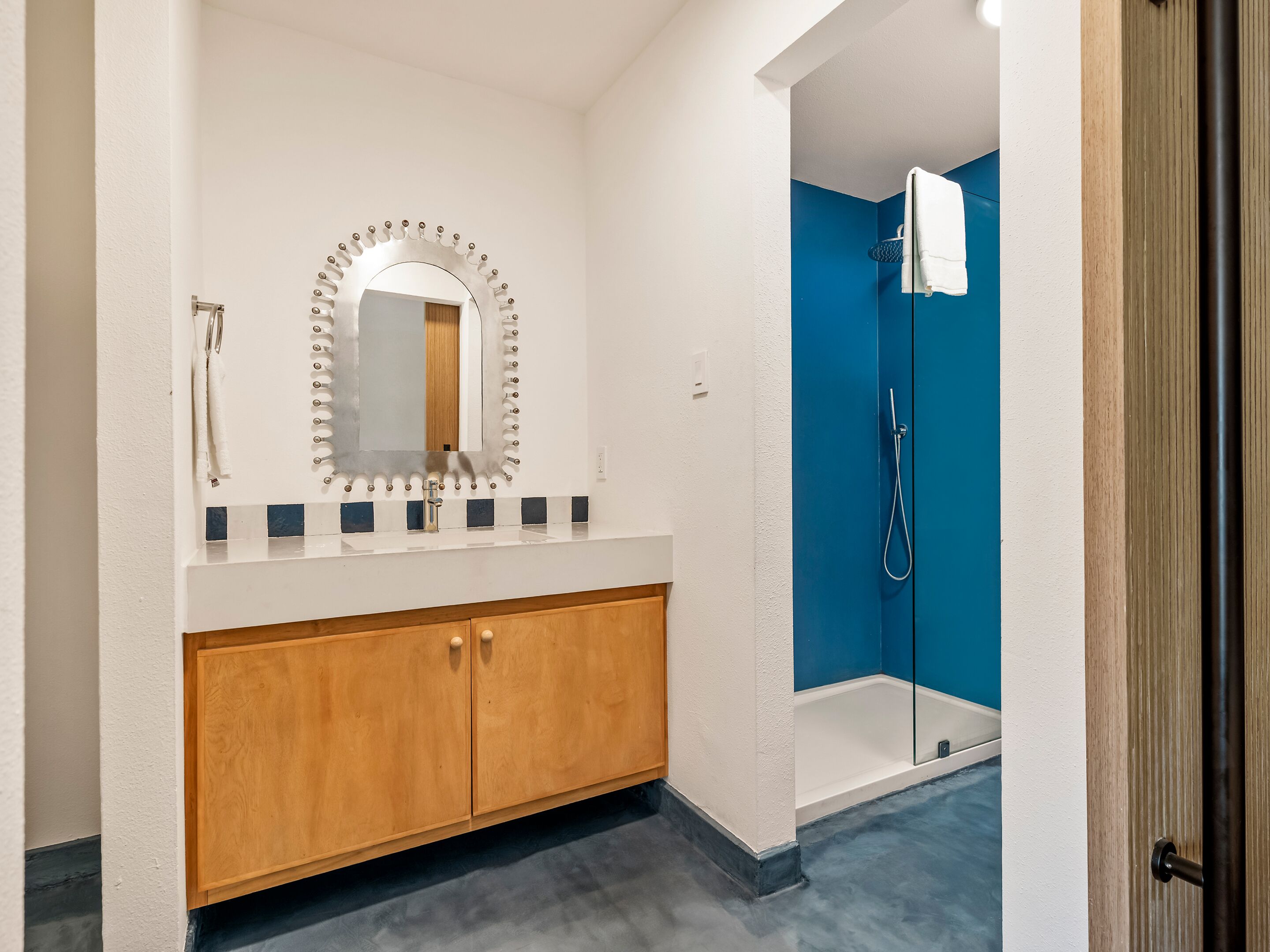 Une salle de bain avec un meuble-lavabo en bois, un grand miroir, une douche avec un mur bleu et une serviette blanche accrochée à la porte.
