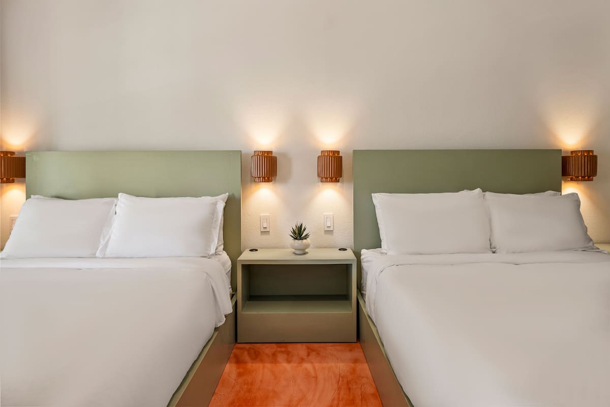 A hotel room with two beds, nightstands, wall-mounted lamps, a small potted plant, and a red carpet.