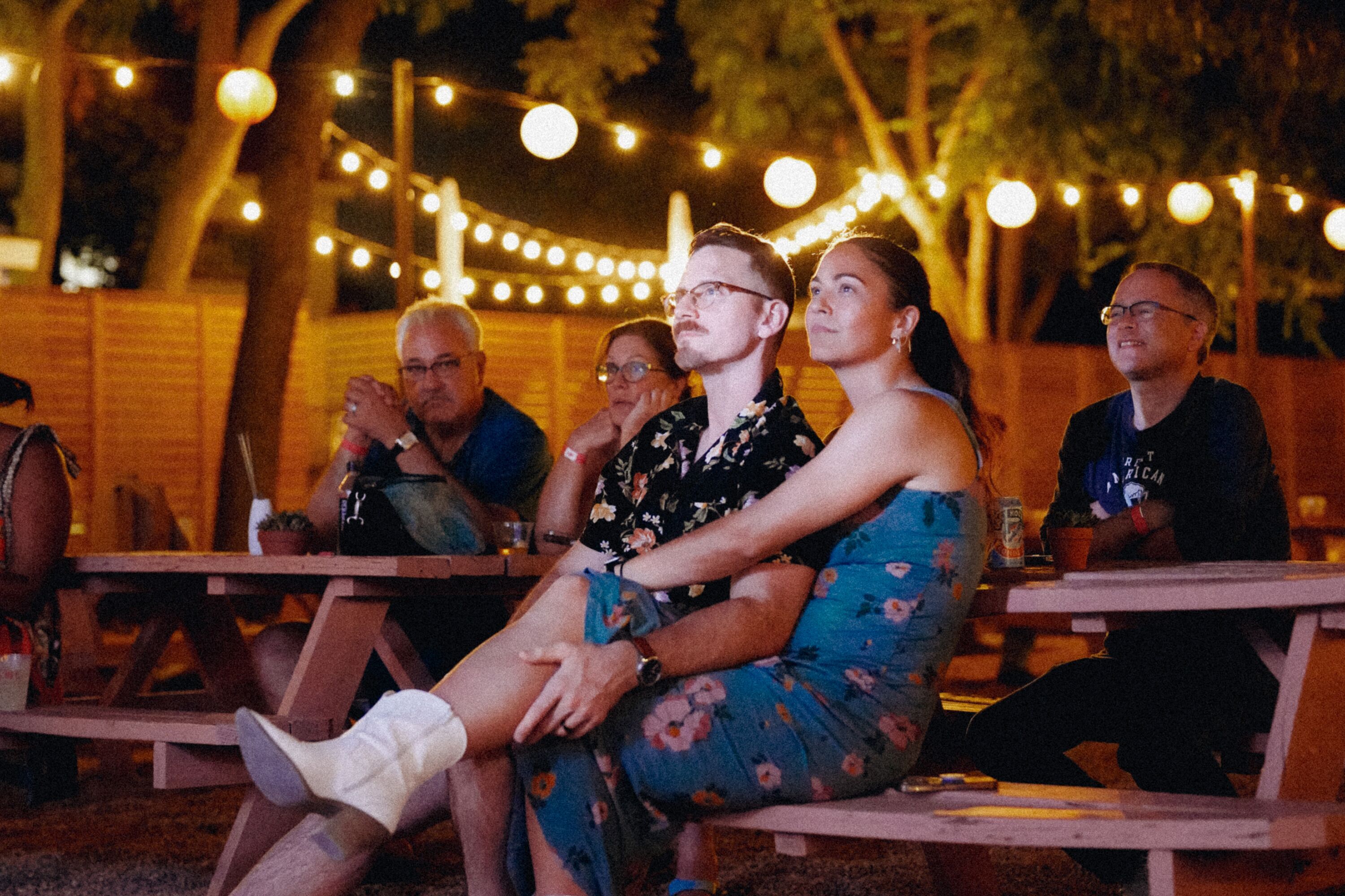 Un groupe de personnes est assis à des tables de pique-nique en bois, à l’extérieur, la nuit, sous des guirlandes lumineuses.