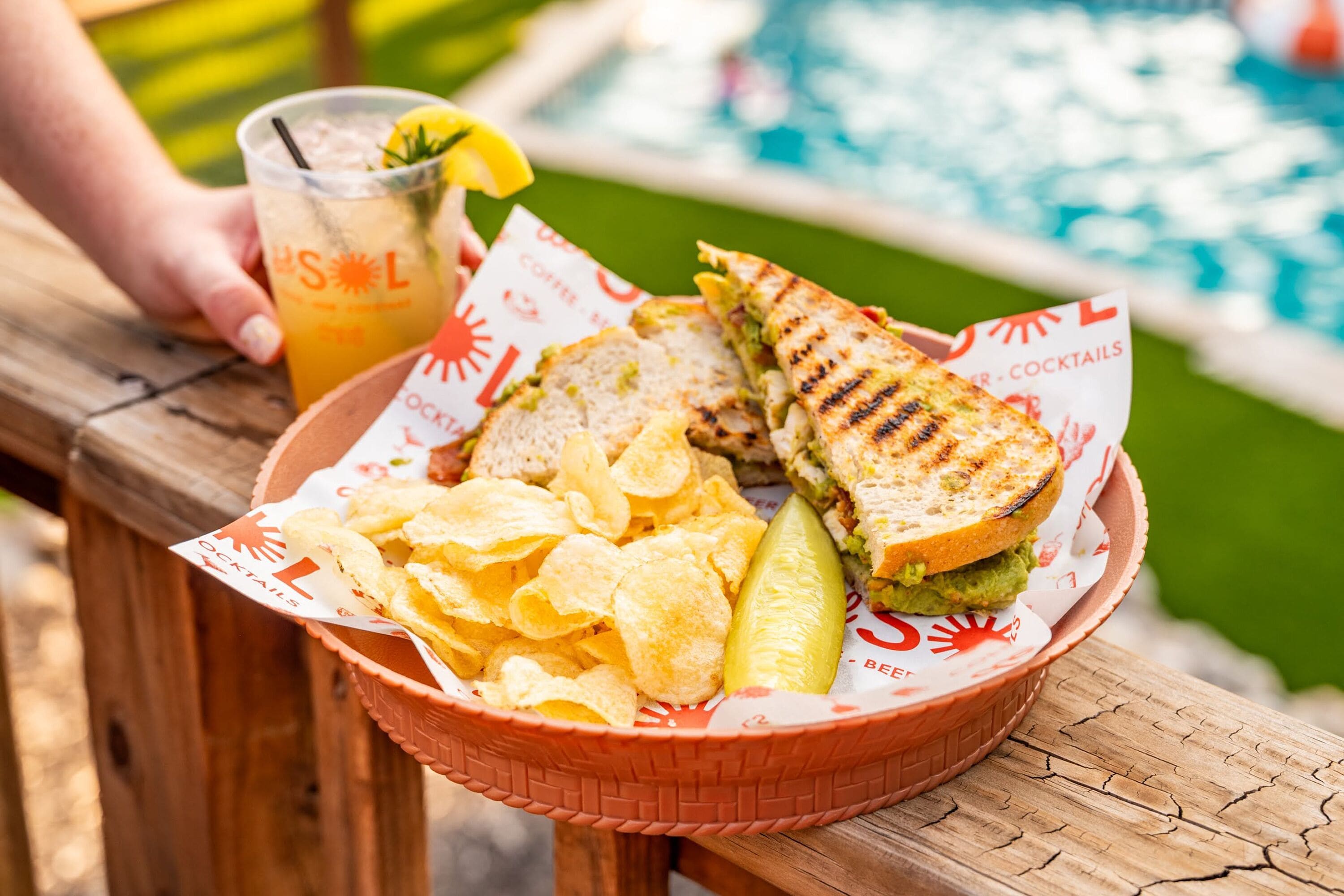 A basket with a grilled sandwich, chips, and a drink on a wooden table near a pool.