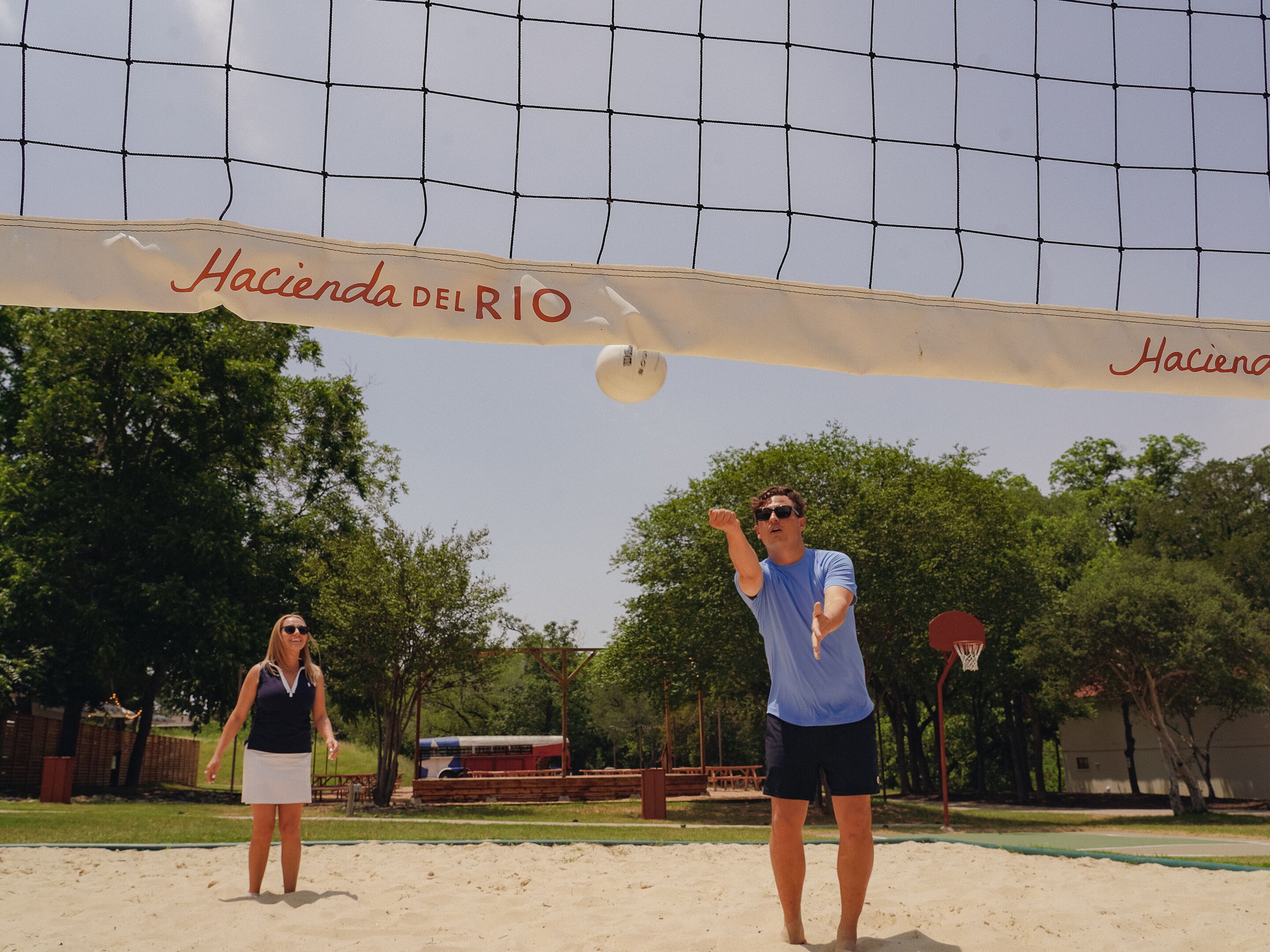 Ein Volleyballnetz mit dem Schriftzug "Hacienda del Rio", aufgestellt in einer sandigen Außenanlage mit Bäumen und einem Basketballkorb im Hintergrund.