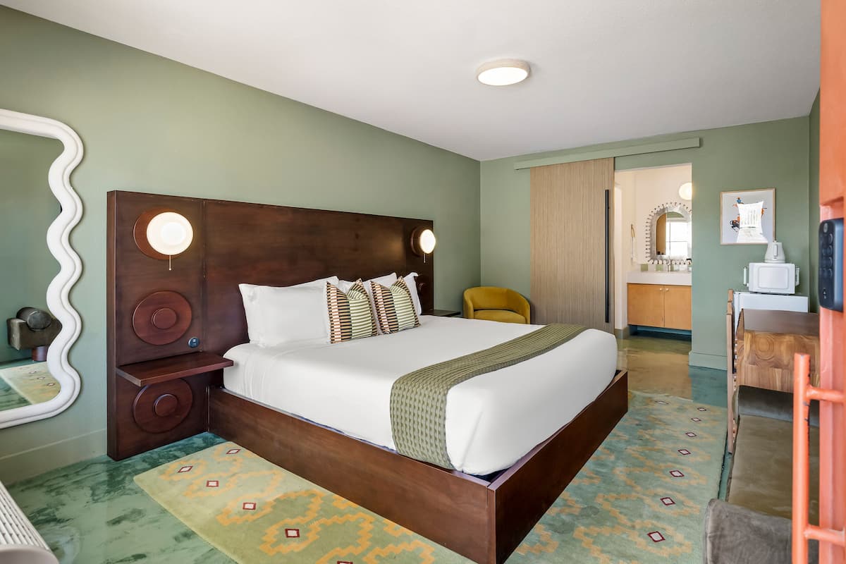 A hotel room with a large bed, a wooden headboard, a mirror, a small table, and a view into the bathroom.