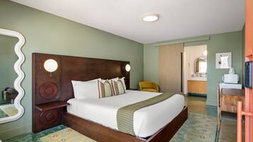 Habitación de hotel con una cama grande, cabecera de madera, un espejo, una mesita y vistas al baño.