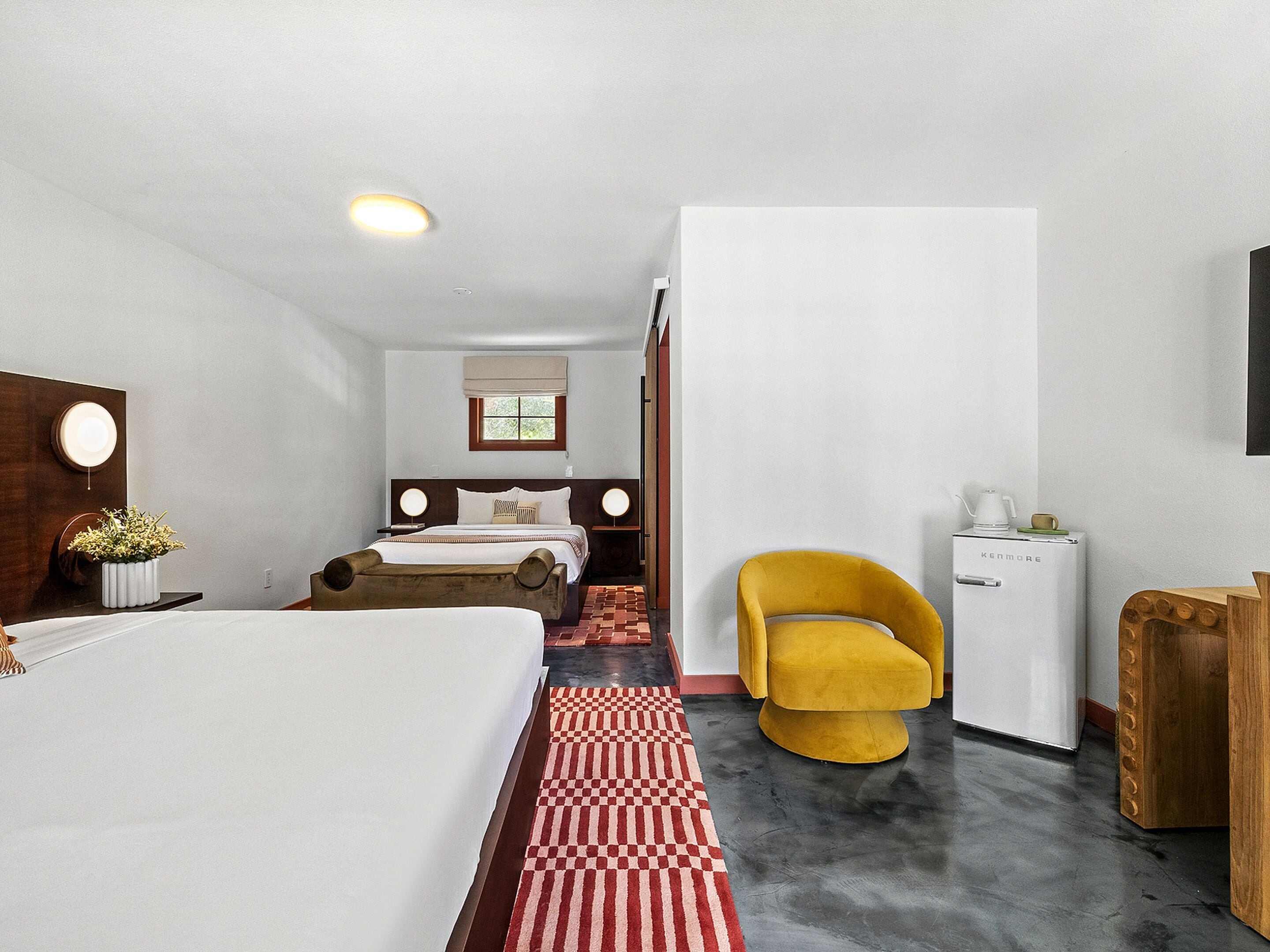 A hotel room with a bed, a yellow chair, a small refrigerator, and a wooden side table.