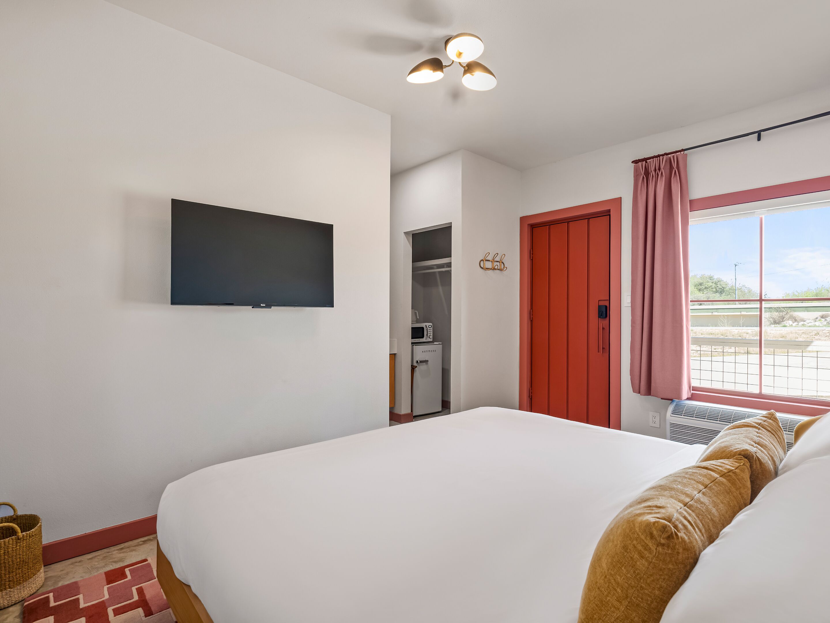 A bedroom with a bed, a flat-screen TV, a microwave, a red door, and a window with curtains.