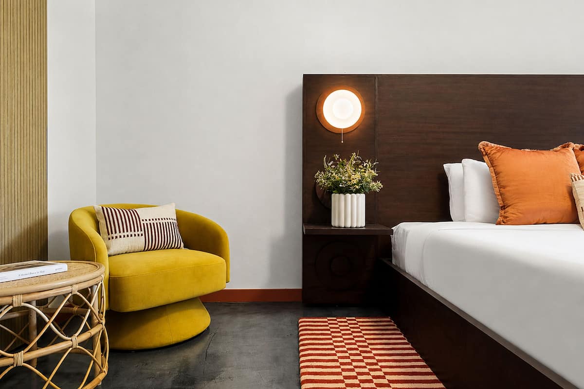A hotel room with a bed, a yellow armchair, a bedside table with a lamp, and a checkered rug.