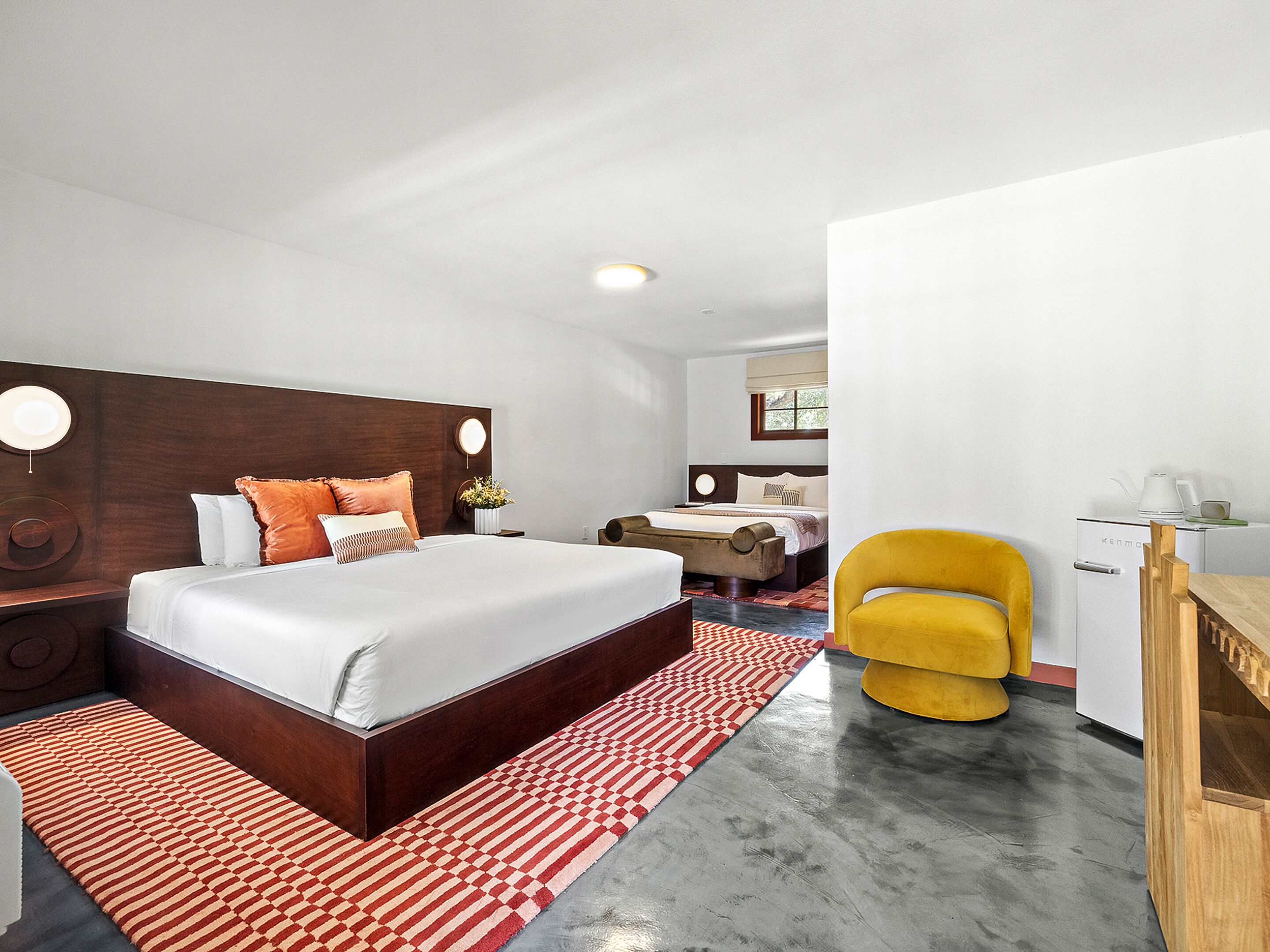 A modern hotel room with a large bed, a desk, a chair, and a kitchenette.