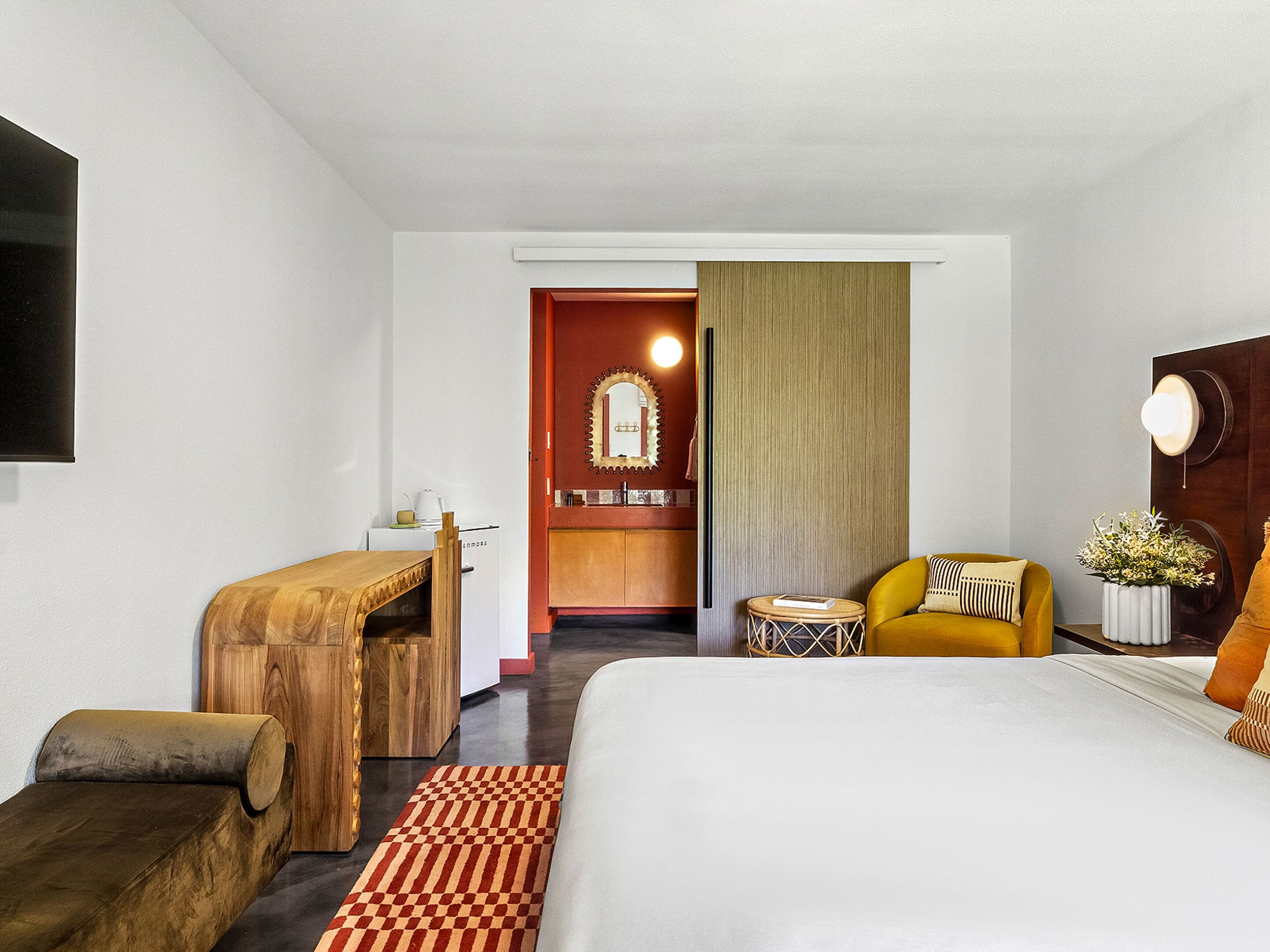A hotel room with a bed, a sofa, a small table, a chair, a mirror, and a view into the kitchen area.