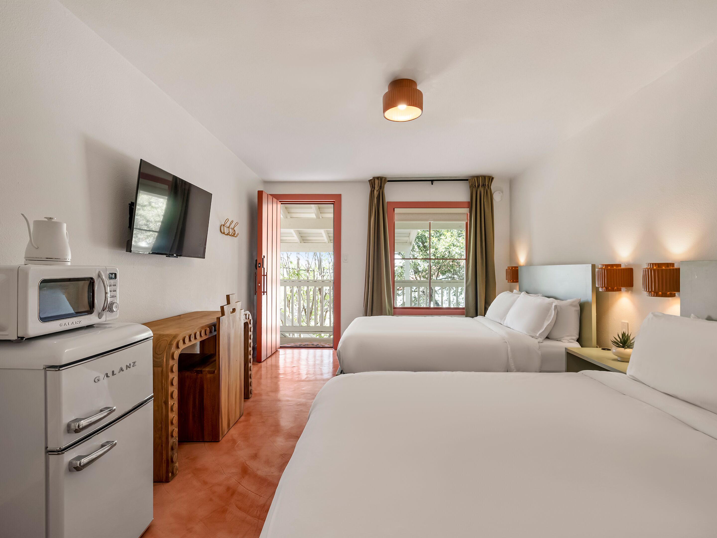 A hotel room with a bed, a desk with a microwave, a TV, a window with curtains, and a ceiling light.