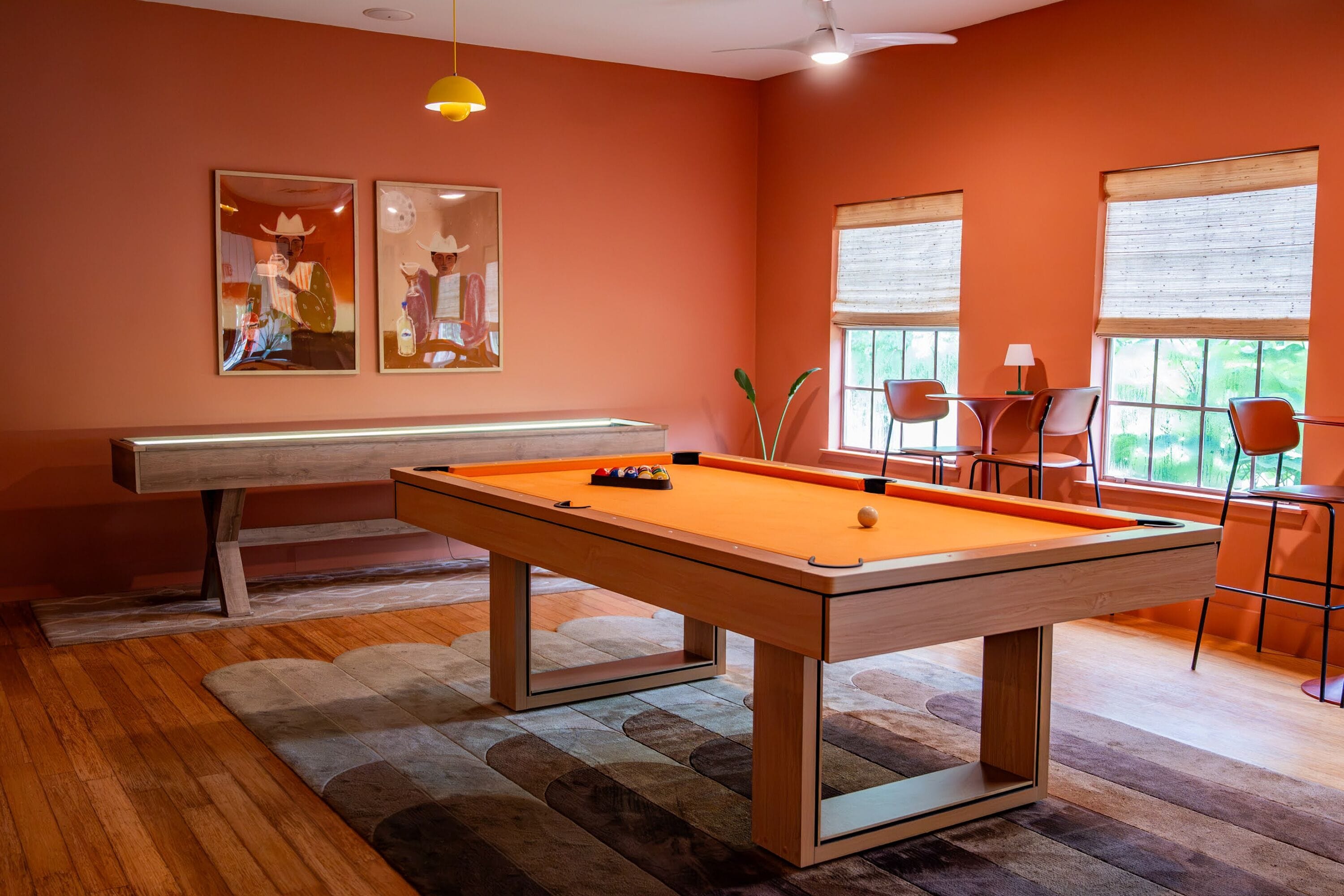 A pool table with a wooden frame, a billiards cue, and a billiard ball. There are two framed artworks on the wall, a ceiling fan, and a window with blinds.