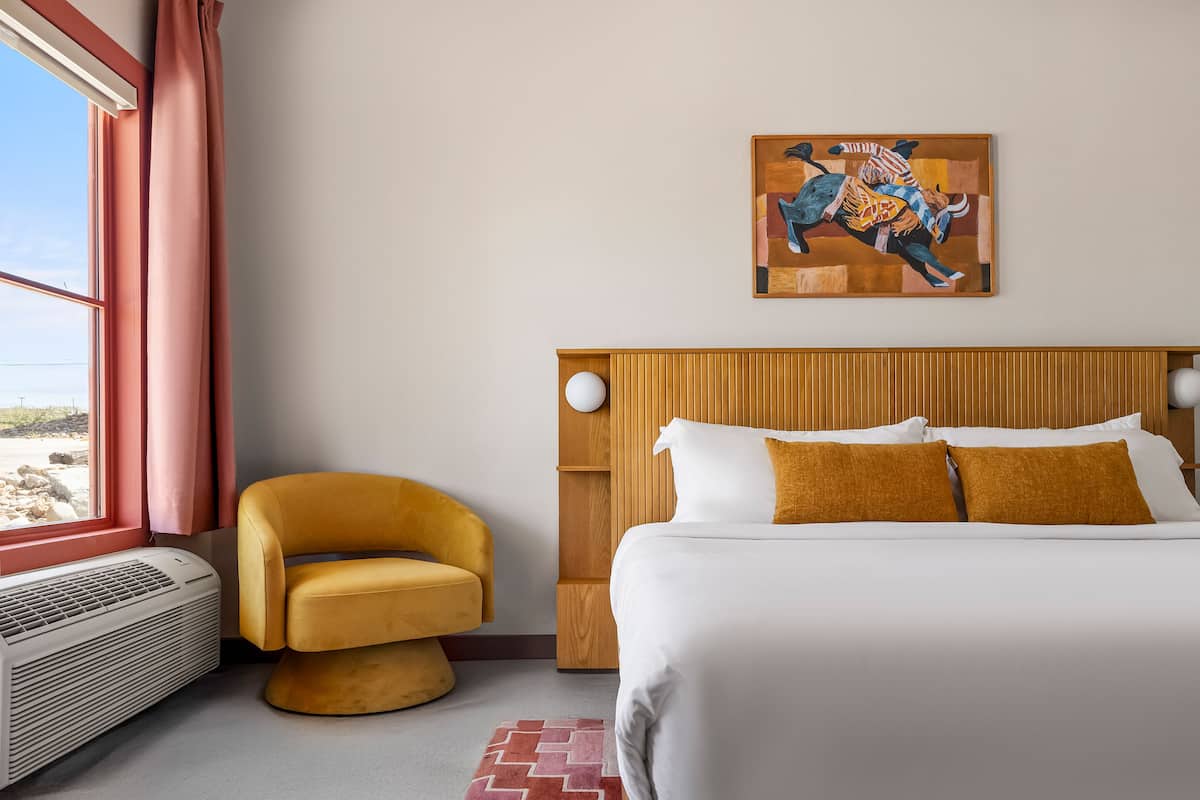 A hotel room with a bed, a chair, a painting, and a window with curtains.