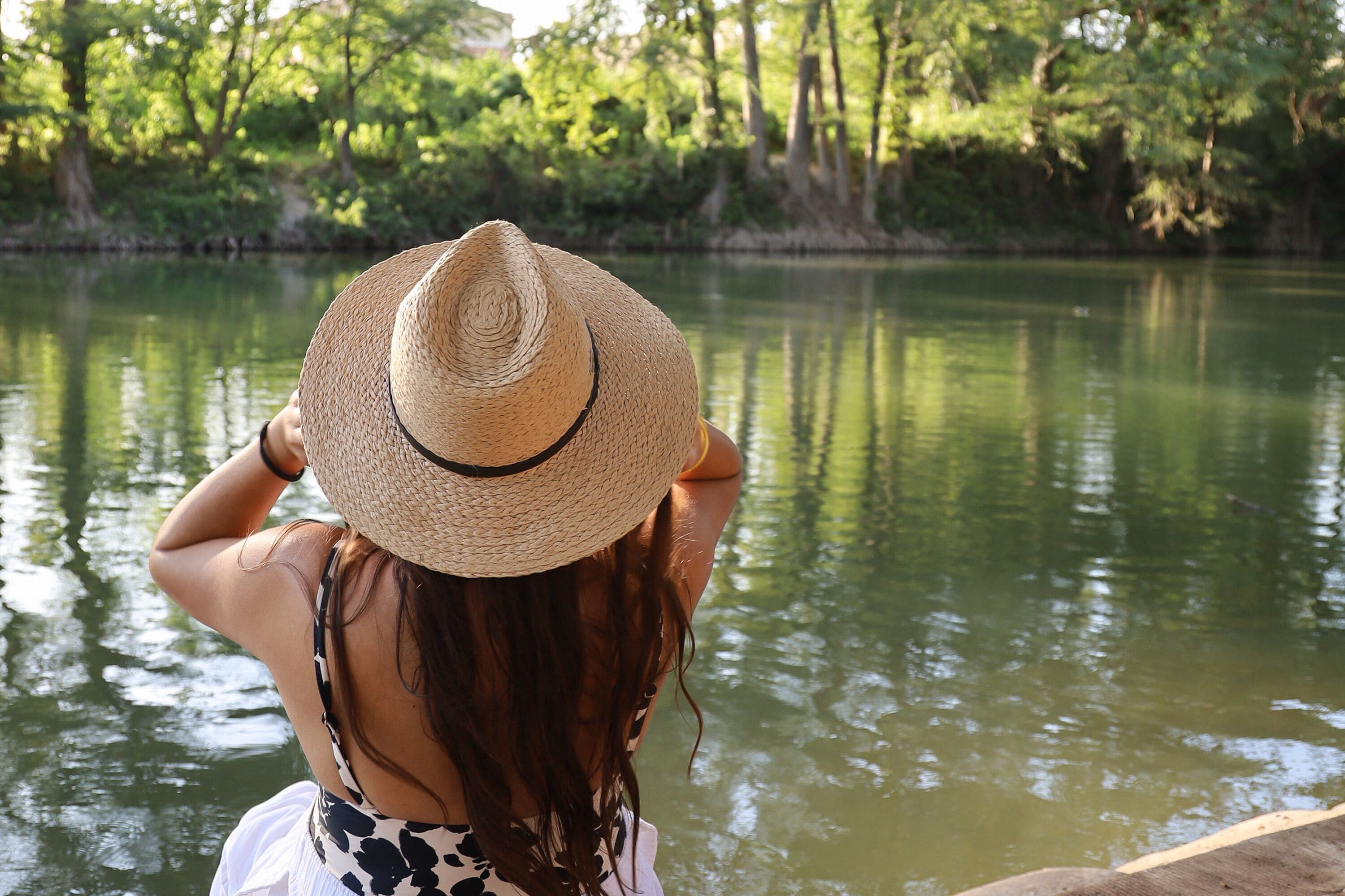 Une personne, vue de dos, portant un chapeau de paille, se tient près d’une étendue d’eau calme, avec des arbres en arrière-plan.