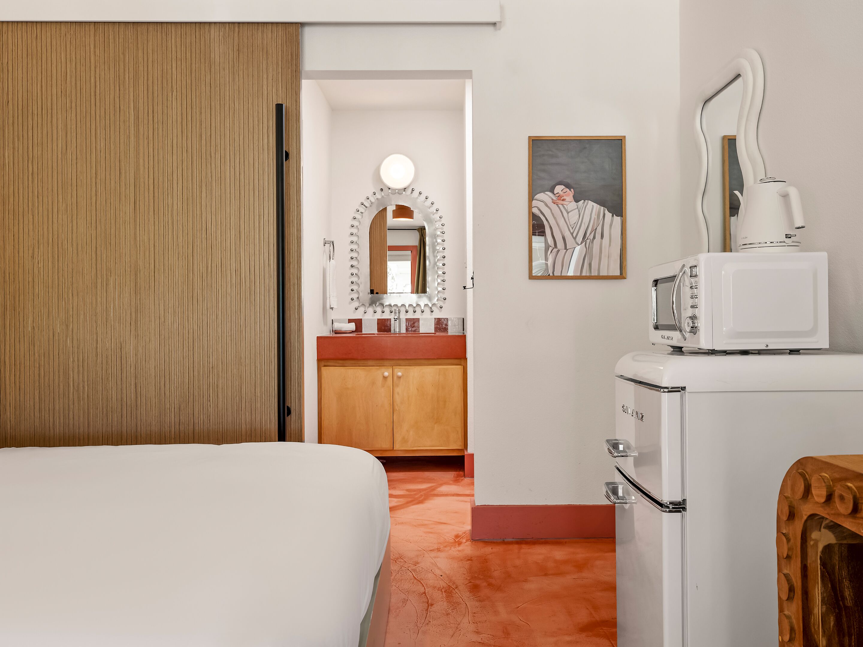 Habitación de hotel con una cama, una puerta de madera, un baño con lavamanos, espejo y toallero.