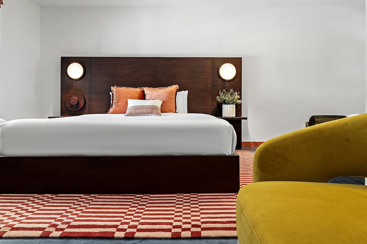 A hotel room with a large bed, a wooden headboard, a small bedside table with a plant, and a yellow armchair.