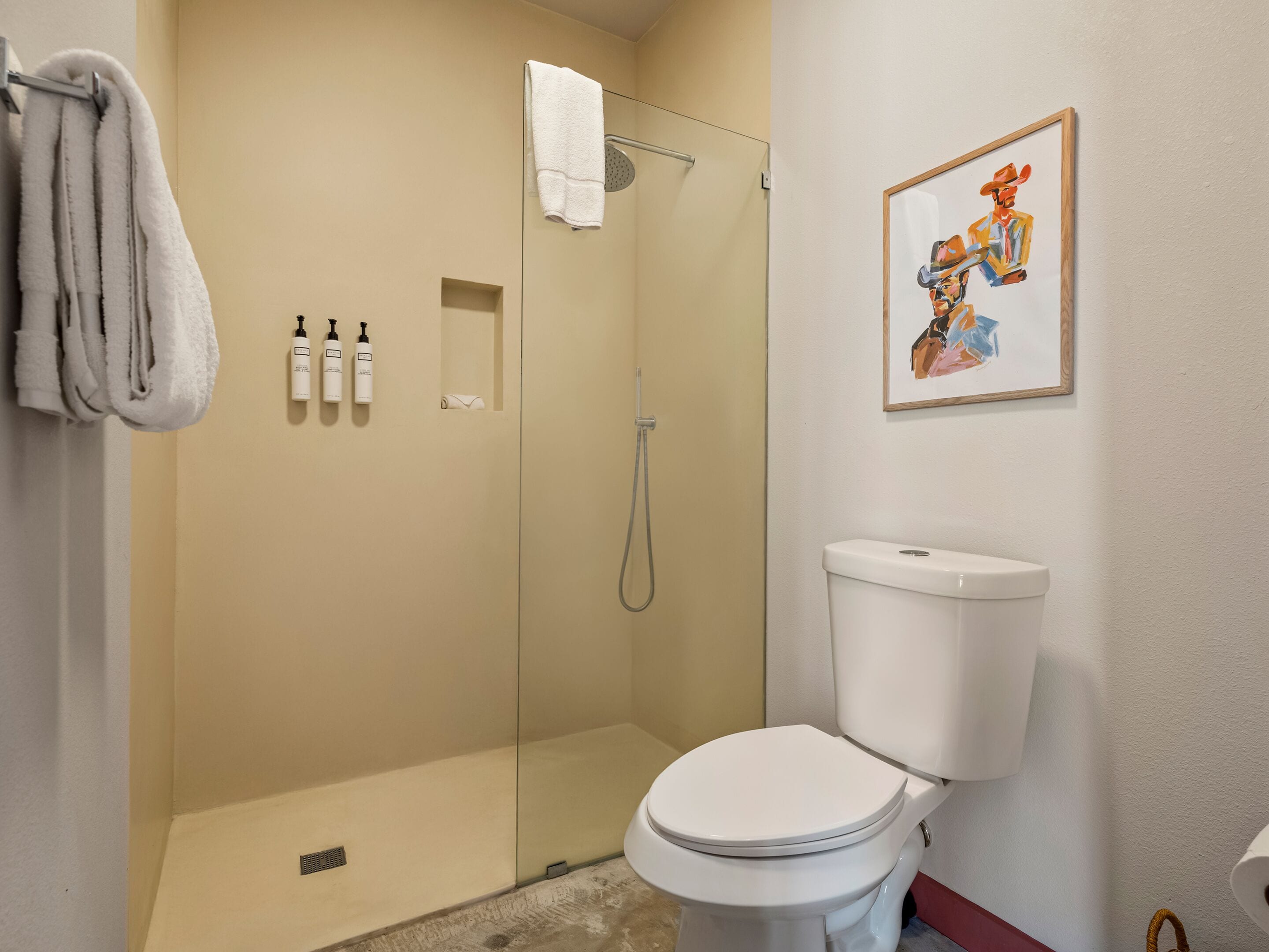 Un baño con un inodoro blanco, una ducha con cerramiento de vidrio y un cuadro en la pared.
