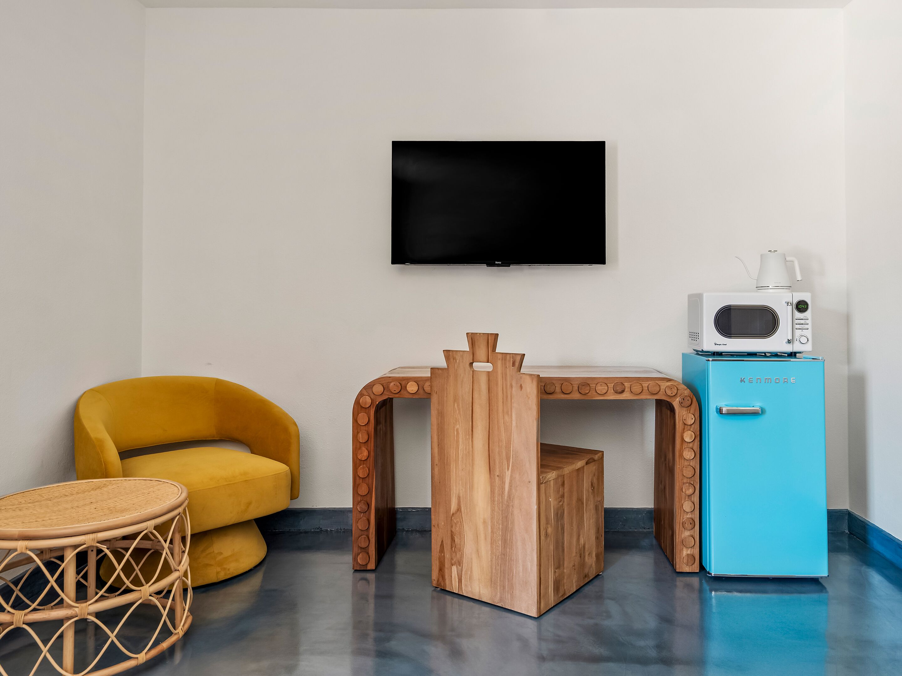 Une pièce avec un fauteuil jaune, une petite table en bois, un réfrigérateur bleu et un téléviseur fixé au mur.