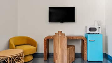 Una habitación con un sillón amarillo, una pequeña mesa de madera, un refrigerador azul y un televisor montado.
