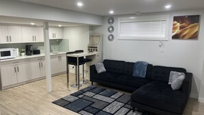 Un espai habitable petit amb una cuineta, un sofà negre i coixins decoratius, i una catifa estampada.