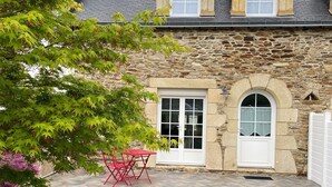 Un bâtiment en pierre avec un toit en pente, une table rouge placée à l’extérieur et une cour couverte d’herbe.