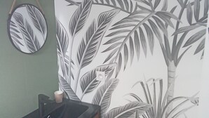 A bathroom with a wooden cabinet, a round mirror, and a wall with a tropical leaf pattern. - La renaissance (Condé-en-Normandie)