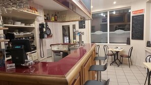 A bar area with stools, a counter, and a coffee machine. There are shelves with bottles and glasses, and a window with a view. - La renaissance (Condé-en-Normandie)