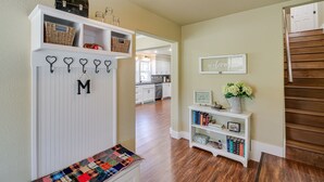 A hallway with a white bench, a wall-mounted quilt, a shelf with baskets and books, and a staircase.