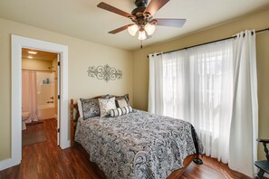 A bedroom with a wooden floor, a bed with patterned bedding, a ceiling fan, and a bathroom visible through an open door. - 'Marie's Place' - Walk to Subiaco Abbey! (Subiaco)