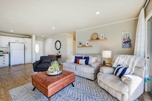 A modern living room with a grey sofa, a brown ottoman, and a black armchair. There is a kitchen in the background with white cabinets and a refrigerator. - Deck, Dock, Fire Pit: Grand Lake Lakefront Home (Jay)