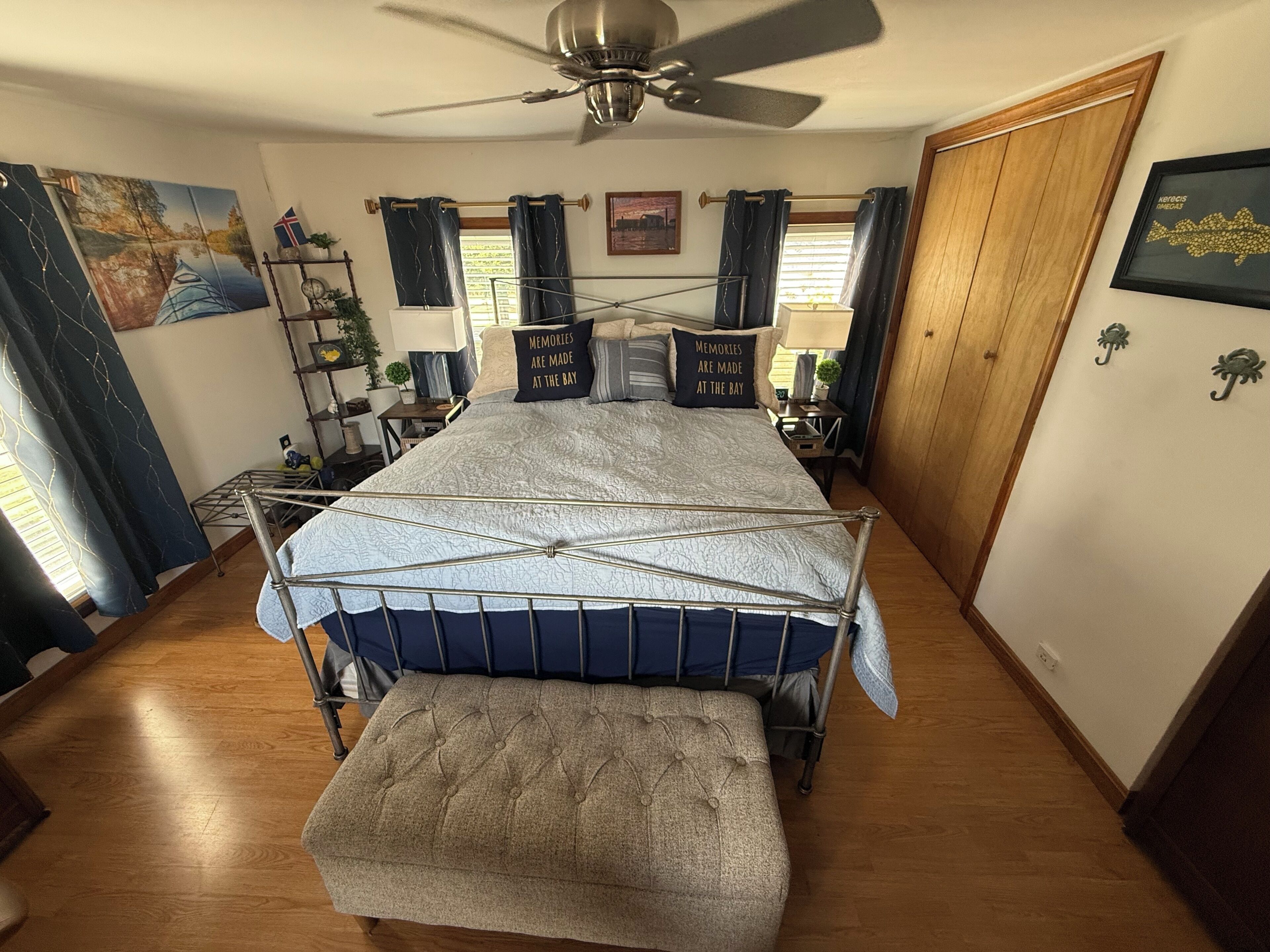 Une chambre à coucher comprenant un lit, un ventilateur de plafond, une table de chevet, une lampe, une étagère avec des objets décoratifs et un tableau accroché au mur.