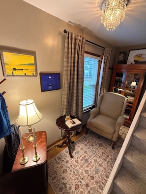 A room with a chair, a lamp, a small table, a rug, a window with curtains, and a chandelier.