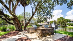 Un patio al aire libre con asientos de piedra y una parrilla, rodeado de árboles y una masa de agua.