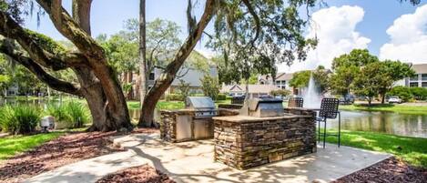 Un patio al aire libre con asientos de piedra y una parrilla, rodeado de árboles y una masa de agua.