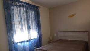 Een slaapkamer met een bed, nachtkastje, lamp en een raam met blauwe gordijnen.