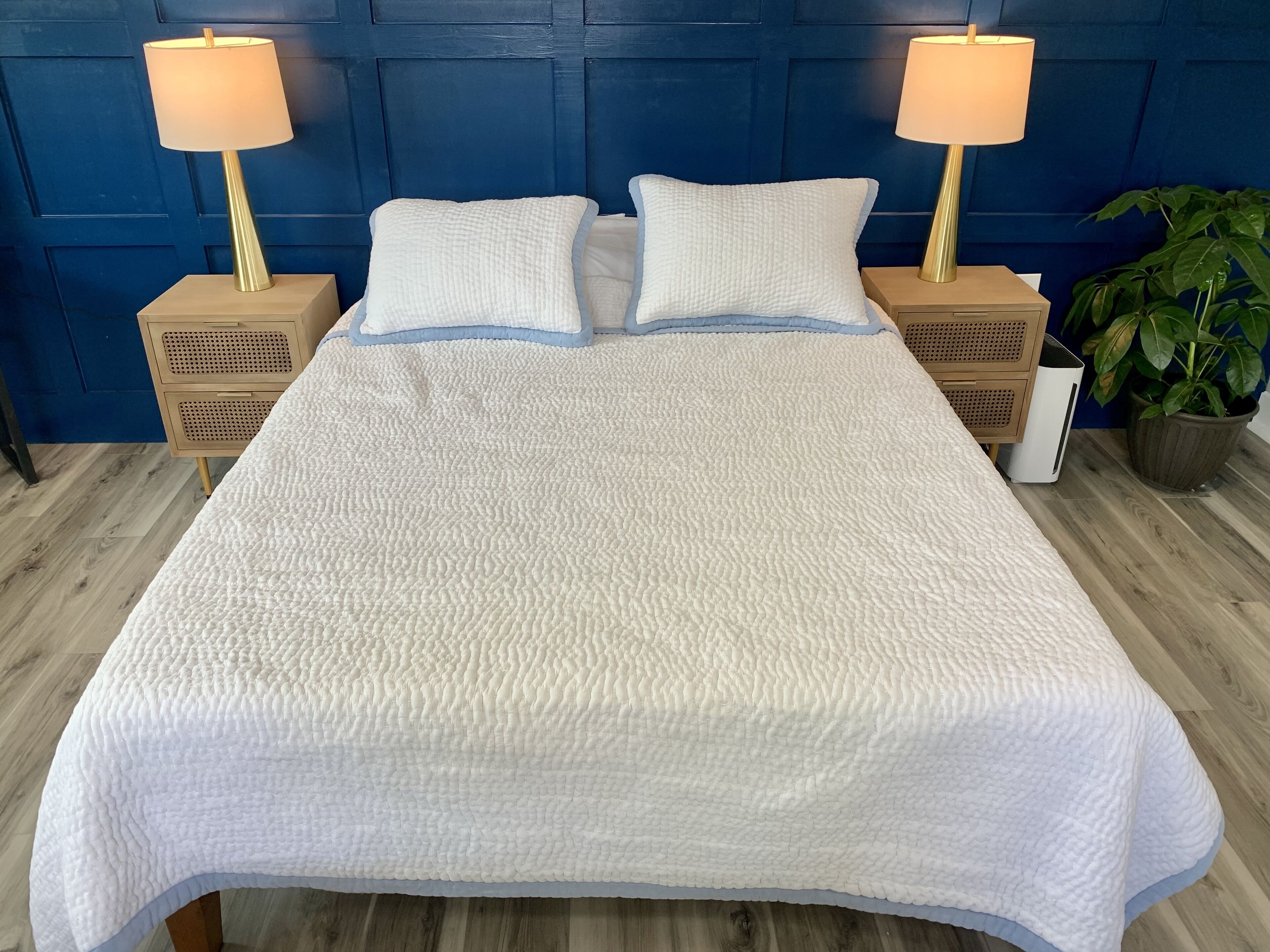 A bed with a textured white cover, two bedside tables with lamps, a blue headboard, and a potted plant.