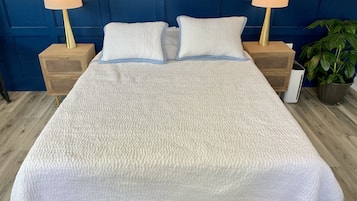 A bed with a textured white cover, two bedside tables with lamps, a blue headboard, and a potted plant.