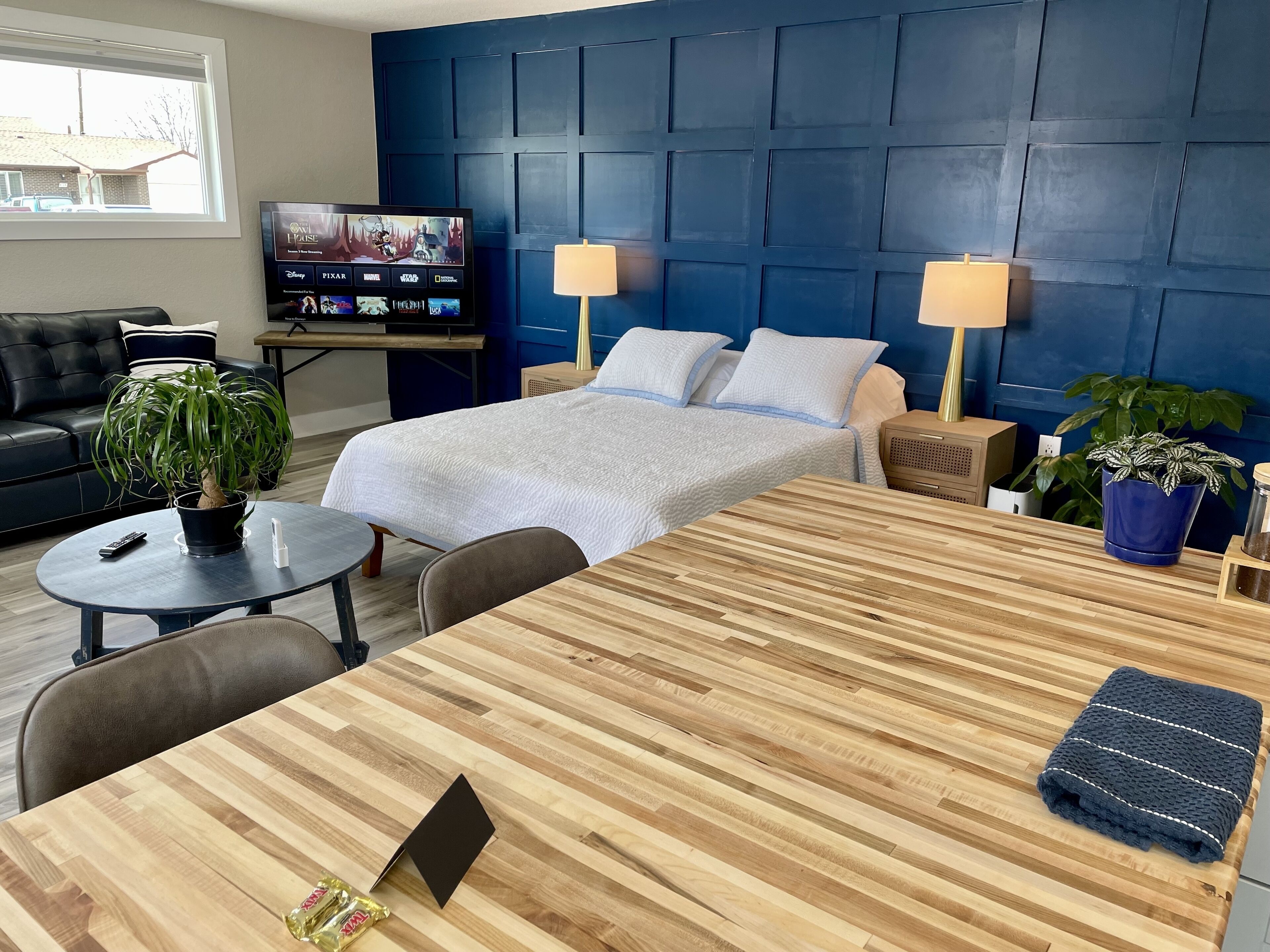 Ein Hotelzimmer mit Bett, zwei Nachttischlampen, einem Fernseher auf einem Tisch, einem Schreibtisch mit Laptop, einem Couchtisch mit Pflanze und einem Blumentopf am Fenster.