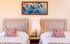 Et hotelværelse med to senge, hver med et matchende sengegærde og pude. Et natbord med en lampe og et lille bord står imellem sengene.