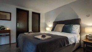 Quarto com cama grande, duas portas, mesa de cabeceira com abajur e uma pequena mesa com uma planta.