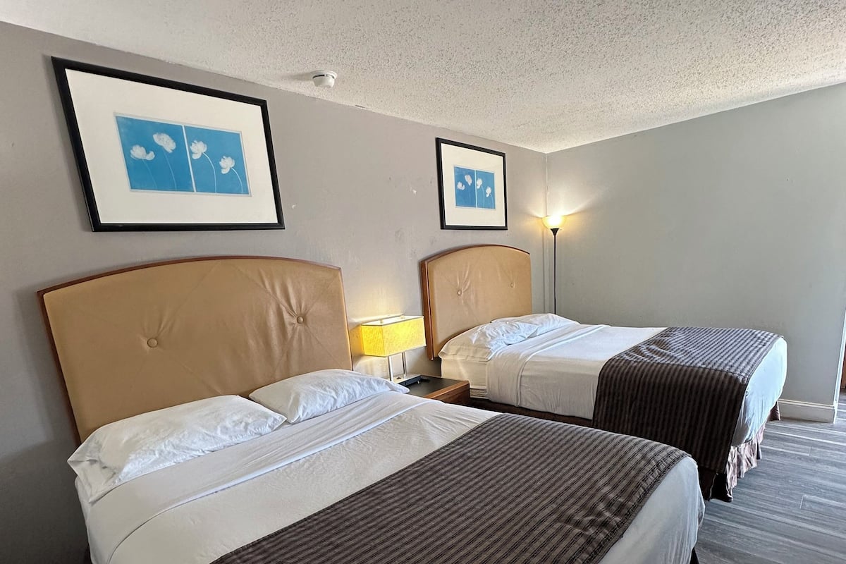 A hotel room with two beds, each with a headboard and a bedside lamp. There are framed pictures on the wall above the beds.