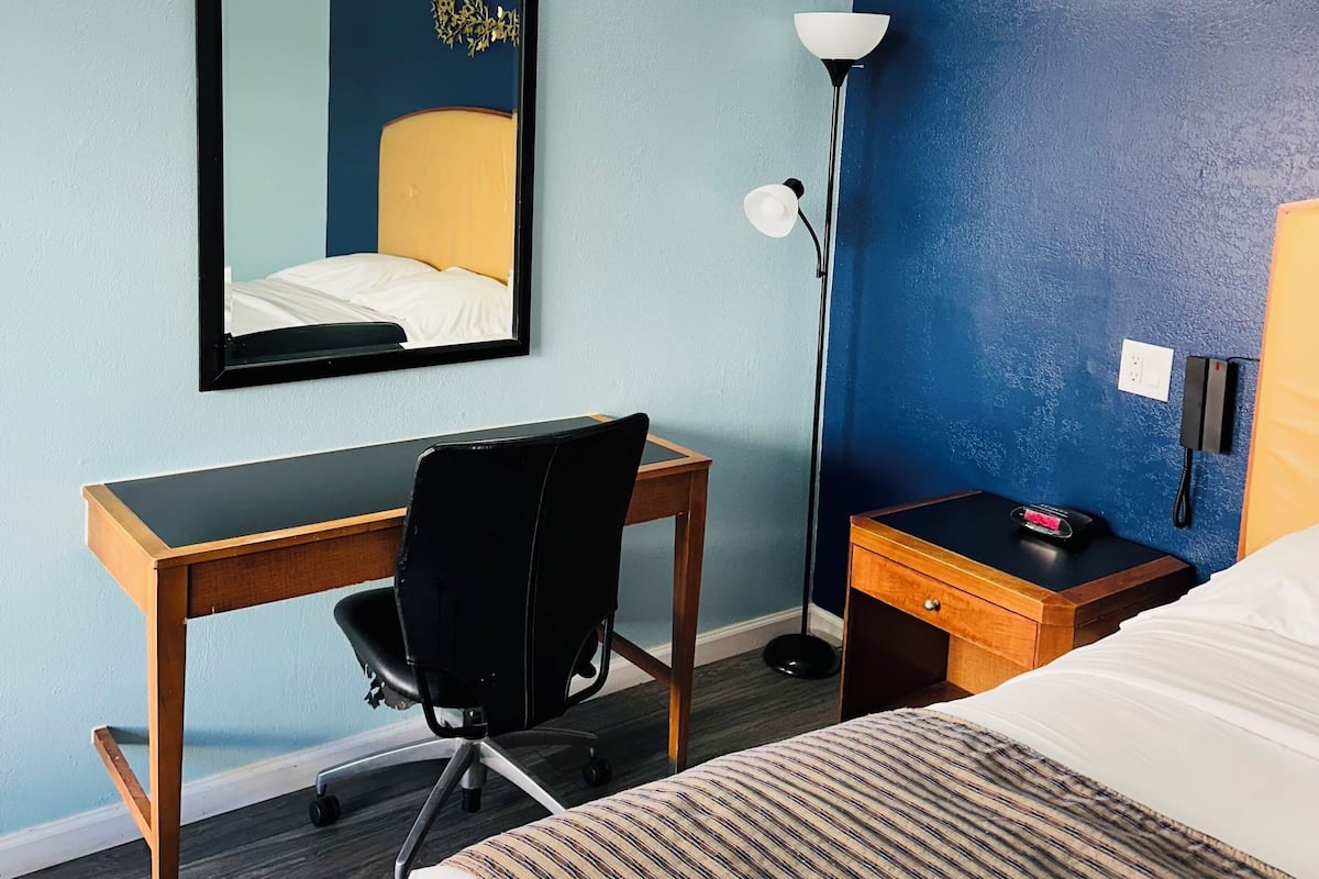 A hotel room with a bed, a desk, a chair, a mirror, a lamp, and a telephone.