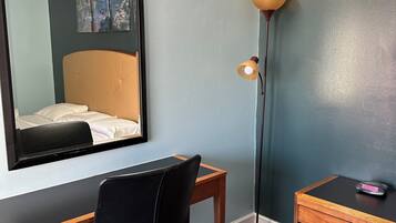 A hotel room with a bed, a desk, a chair, a standing lamp, and a small wooden side table.