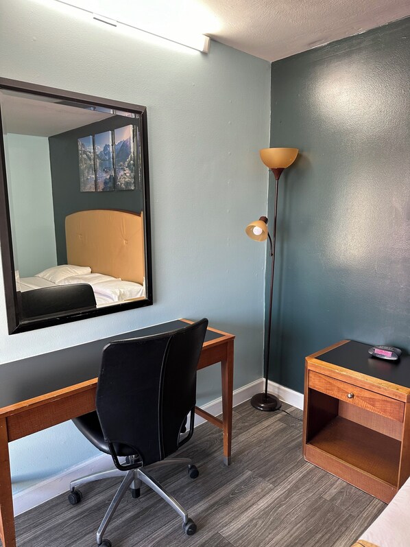 A hotel room with a bed, a desk, a chair, a standing lamp, and a small wooden side table.
