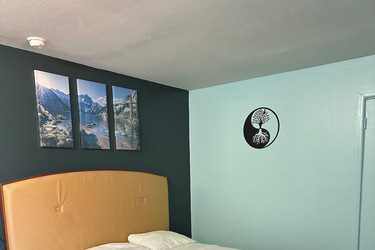A neatly made bed with a headboard, a bedside table, and a wall-mounted picture of mountains.