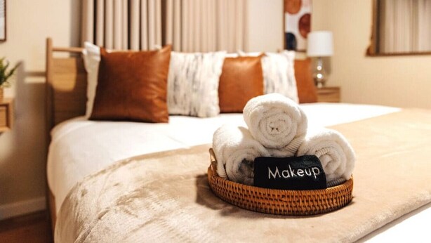 A neatly made bed with a rolled towel in a basket labeled "Makeup" and a brown pillow. - Peachtree/Royal Oak`s Sweet Spot/King Bed (Royal Oak)